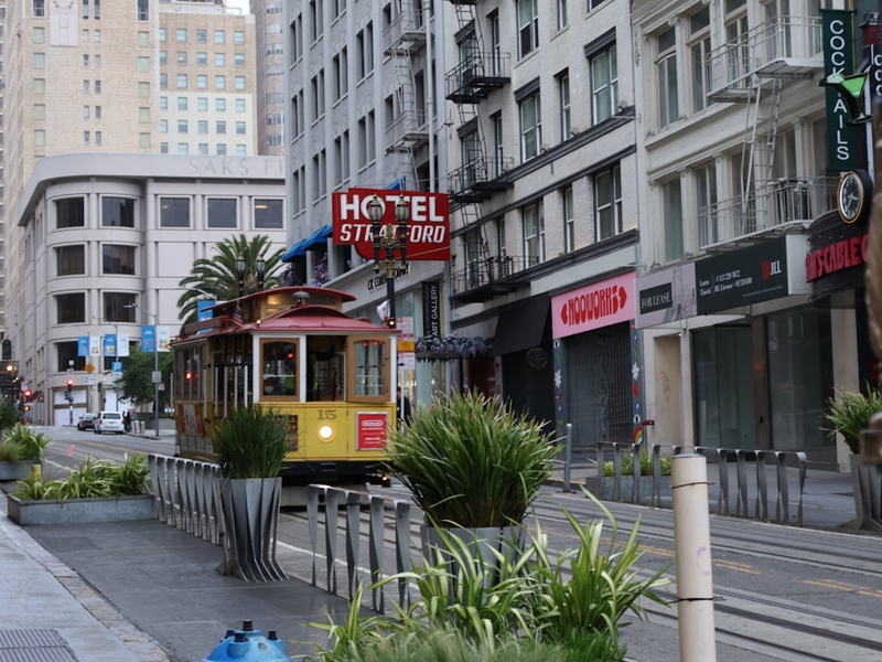 Red and yellow San Francisco cable car passing Hotel St. Ali on a tree-lined urban street with shops, planters, and high-rise buildings under an overcast sky in California.