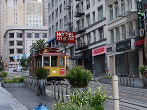 Red and yellow San Francisco cable car passing Hotel St. Ali on a tree-lined urban street with shops, planters, and high-rise buildings under an overcast sky in California.