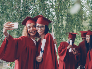 Pretty female graduates are taking selfie with diploma scroll using smartphone, young women are posing with other students in gowns moving and talking in background.