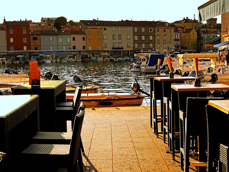 Outdoor cafe tables by a waterfront with buildings.