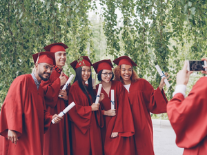 Multiethnic group of graduating students is posing for smartphone camera, waving hands with diplomas and shouting while guy is shooting them touching screen and gesturing.