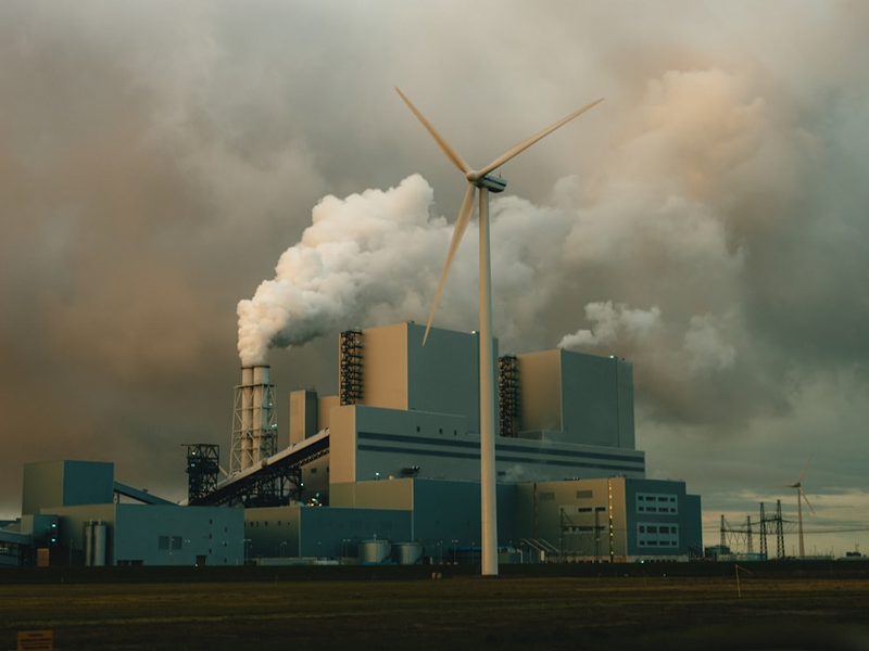 In the heart of Eemshaven's industrial harbor, a lone wind turbine stands tall, asserting its presence beneath the stormy skies of the Netherlands. As the sun dips below the horizon, casting an orange glow, a coal-burning power station looms in the background, with thick smoke billowing into the air.
