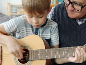 Caring grandfather is helping cute kid teaching guitar playing at home together enjoying domestic music lesson. People and musical education concept.