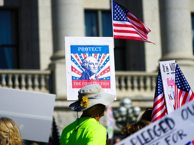 Protestors hold signs and american flags outside building.