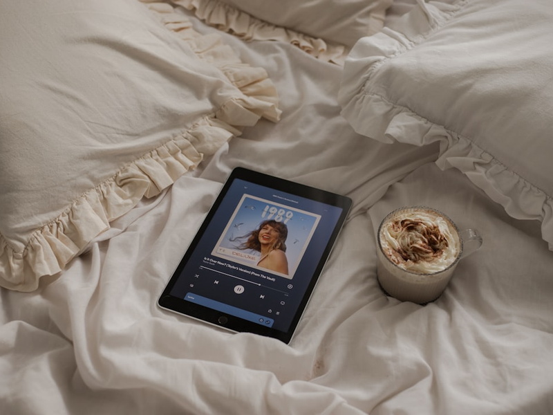 An iPad 9th generation playing "Is It Over Now? (Taylor's Version) (From The Vault)" by Taylor Swift on Spotify lying on the white bed sheets next to a mug of hot chocolate || 📸 Photo by: Jovan Vasiljević