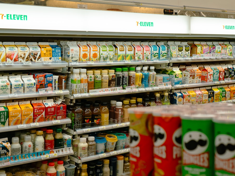 A brightly lit refrigerated aisle in a Taiwanese 7-Eleven showcases an immense variety of colorful beverage cartons and bottles. Neatly organized shelves are packed with everything from fresh milk and soy milk to teas and juices, reflecting the central role of convenience in modern East Asian urban life. The vibrant packaging creates a dazzling mosaic under the cool, fluorescent glow. In the foreground, the familiar shape of Pringles cans adds a touch of global branding to the local selection, creating a sense of abundant choice.