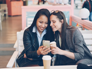 Pretty young ladies friends are using modern smartphone sitting at table in cafe with take away coffee. Happy girls are touching screen, watching funny photos and laughing.