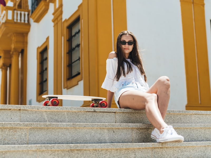 a stylish young student girl in sunglasses sits on the steps and next to her is a skateboard.