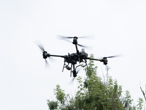 A powerful industrial drone hovers above treetops against a pale sky, caught mid-flight while delivering solar panels—showcasing the cutting edge of aerial logistics and clean energy deployment.