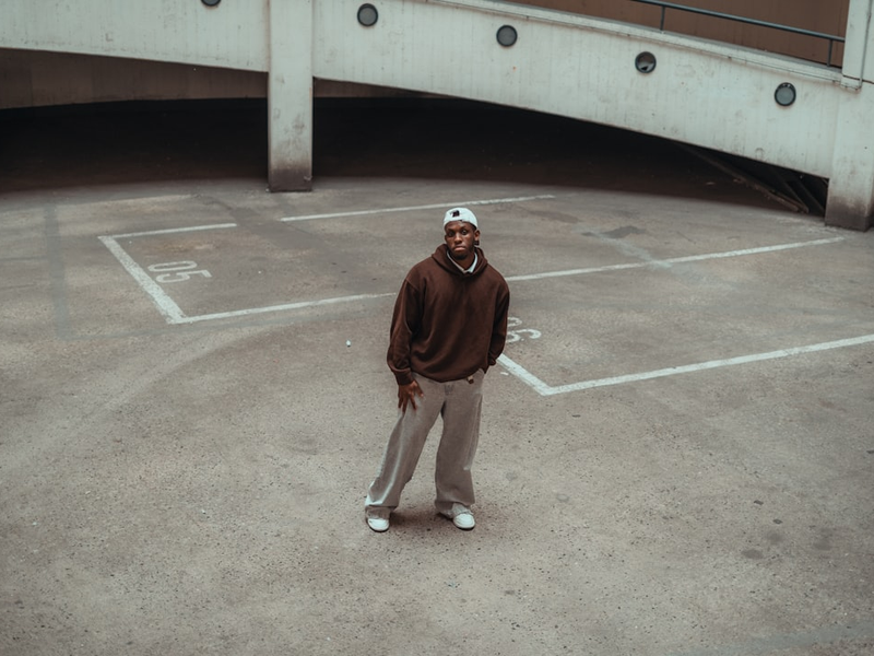 A wide-angle fashion shot of a man standing in the center of an empty concrete parking garage. He is wearing a brown hoodie, baggy grey trousers, and a white cap, showcasing a relaxed streetwear aesthetic against an industrial architectural backdrop.