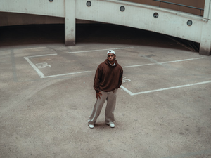 A wide-angle fashion shot of a man standing in the center of an empty concrete parking garage. He is wearing a brown hoodie, baggy grey trousers, and a white cap, showcasing a relaxed streetwear aesthetic against an industrial architectural backdrop.