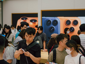 A bustling crowd fills the Apple Store, generating a buzz of excitement on the highly anticipated launch day of the iPhone 17 series. Customers lean over the signature wooden display tables to get their first hands-on experience with the new devices, while massive wall screens showcase the sleek design and triple-lens camera system of the latest iPhone 17 Pro.