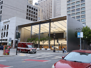 Iconic Apple Store at Union Square in San Francisco, California, with its distinctive glass cube facade amid skyscrapers, traffic, an ambulance, and pedestrians on a cloudy urban day.