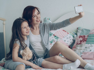 Happy mother and little girl taking selfie photo with smartphone camera and have fun grimacing while sitting in bright bedroom at home. Family, people and technology concept