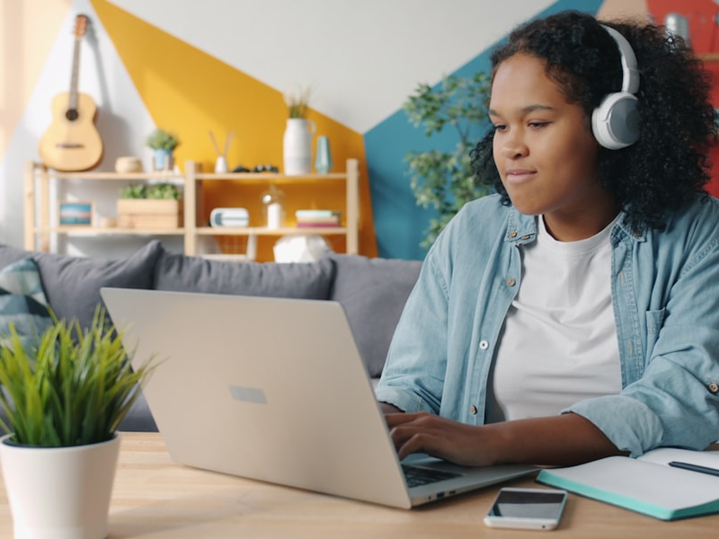 Joyful Afro-American female student in headphones is using laptop at home dancing having fun alone. Modern technology, youth and emotions concept.