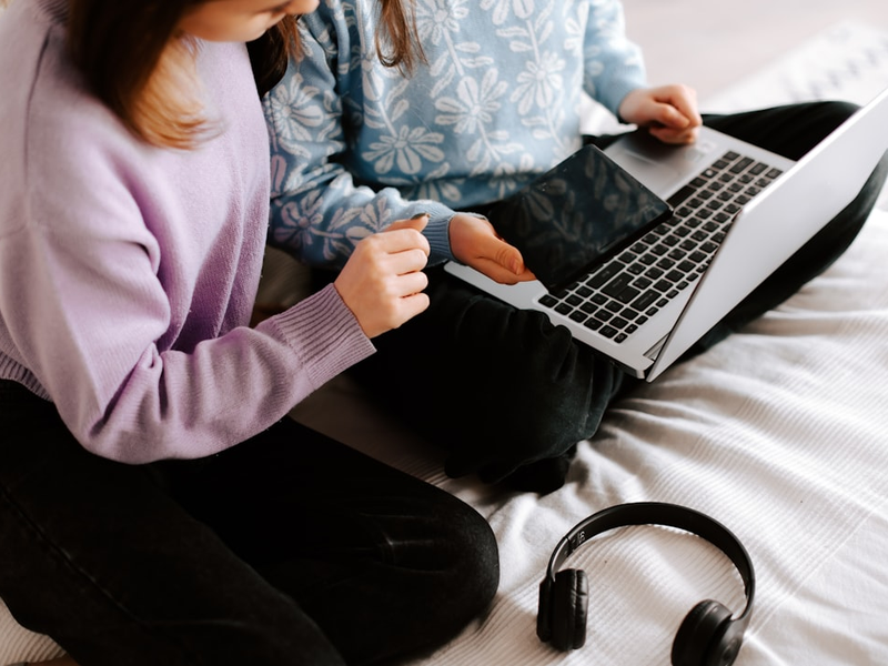 two girls sitting on a bed with a laptop and headphones