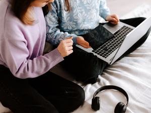 two girls sitting on a bed with a laptop and headphones