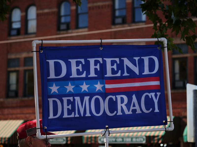 Sign reads "defend democracy" with american flag elements.