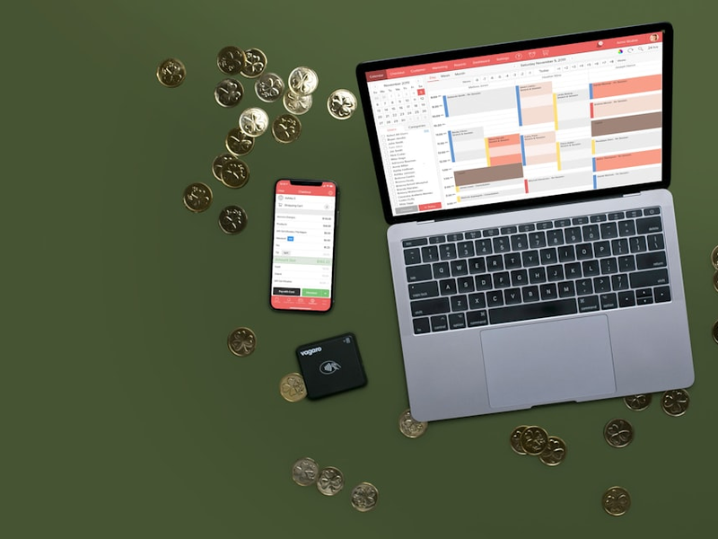 On a dark green background sits a laptop, phone, and credit card reader with gold coins scattered around it. the Laptop has a booking calendar displayed while the phone has a checkout screen.
