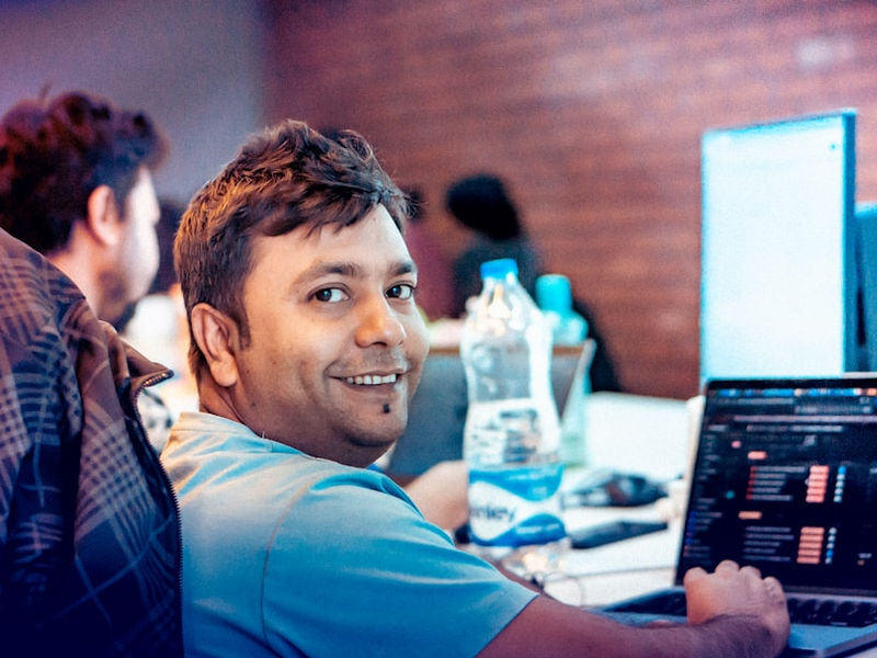 A cheerful man smiles warmly while working on his laptop in a modern office setting. The glowing computer screen and soft background blur create a vibrant workplace vibe, perfect for showcasing positivity, teamwork, and digital work culture.