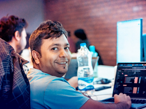 A cheerful man smiles warmly while working on his laptop in a modern office setting. The glowing computer screen and soft background blur create a vibrant workplace vibe, perfect for showcasing positivity, teamwork, and digital work culture.