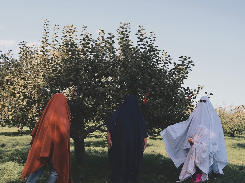 Three people in ghost costumes stand near a tree.