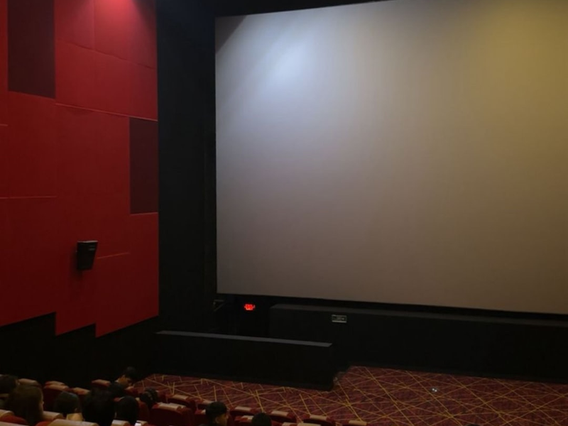 A modern cinema lobby interior featuring vibrant red and white ceiling lighting, digital menu boards, a food counter, and people waiting for tickets. The atmosphere is sleek and contemporary, with a shiny black tiled floor and unique architectural elements, including wooden pillars and artistic lighting design.