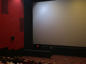 A modern cinema lobby interior featuring vibrant red and white ceiling lighting, digital menu boards, a food counter, and people waiting for tickets. The atmosphere is sleek and contemporary, with a shiny black tiled floor and unique architectural elements, including wooden pillars and artistic lighting design.
