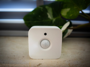 This image features a Philips Hue motion sensor, designed to seamlessly integrate into smart home ecosystems. The compact, sleek white square device with a prominent sensor lens is shown resting on a natural stone surface with greenery and window blinds softly blurred in the background. The photo conveys a modern, clean, and tech-friendly atmosphere, ideal for illustrating smart home technology, automation setups, or home security concepts. The soft lighting enhances the minimalist and functional design appeal of the device.