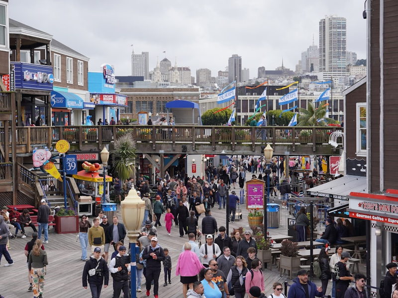 A bustling crowd fills the wooden boardwalk of Pier 39, surrounded by colorful storefronts, iconic signage, and layers of decks lined with flags and flowers. The San Francisco skyline rises in the background under a moody gray sky, adding a dramatic urban contrast. Shops like Trish’s Mini Donuts and Aquarium of the Bay create a lively, family-friendly atmosphere.