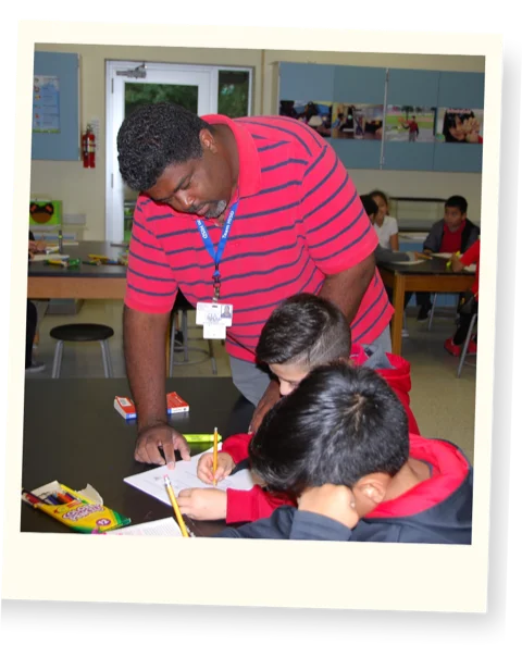Alex Jones leaning over a desk to help two elementary students with a science worksheet