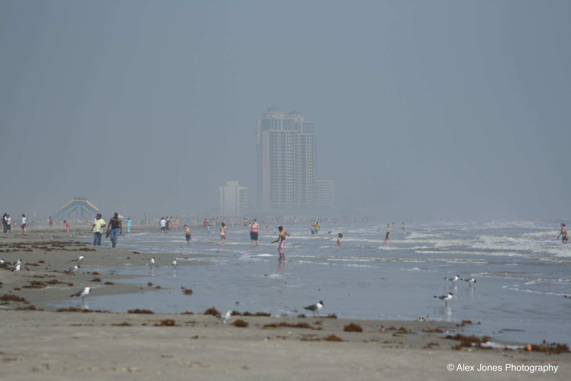 This beach scene shows people playing in the ocean water on a foggy day. A tall building in the background is partly hidden by thick fog or mist.