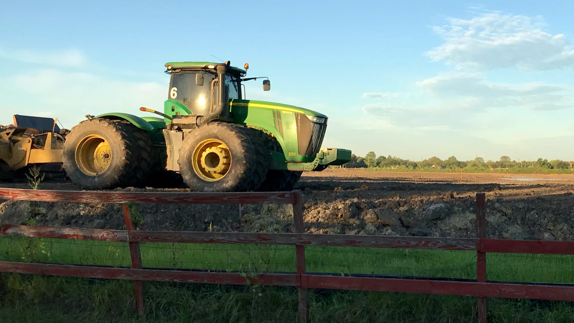 A big green tractor with huge wheels is working in a dirt field. The tractor is pulling farm equipment to prepare the soil for planting crops.