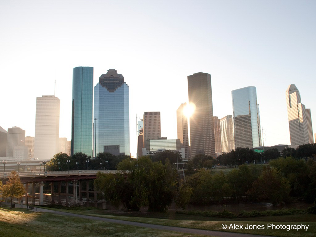 This picture shows tall buildings in a city during sunrise or sunset. The sun is shining brightly between the buildings, creating shadows and light.