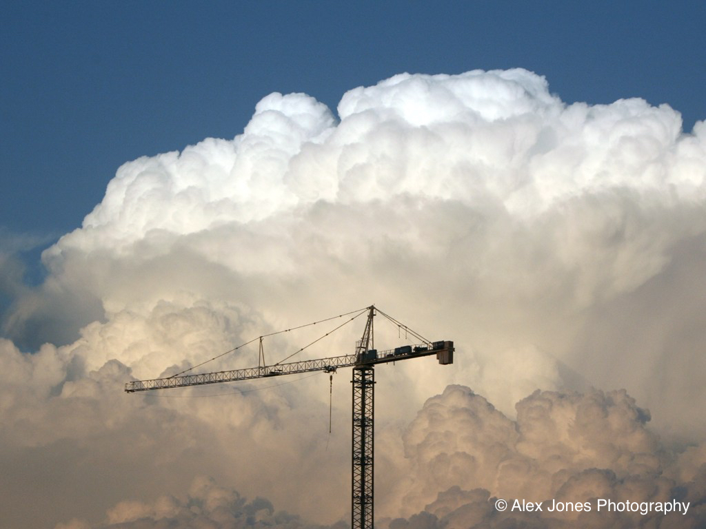 A tall construction crane stands against a sky filled with big, puffy white clouds.