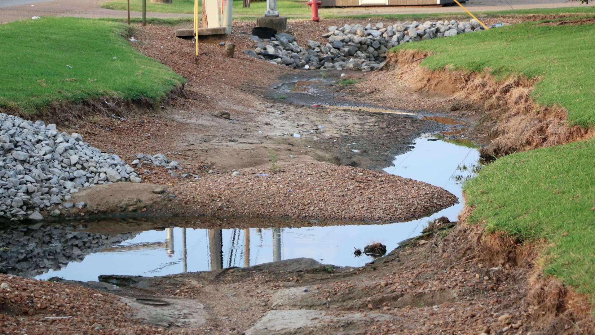 The image shows a channel with a small stream of water flowing through it. The banks of the channel are steep and show signs of erosion, with exposed soil and grass roots.