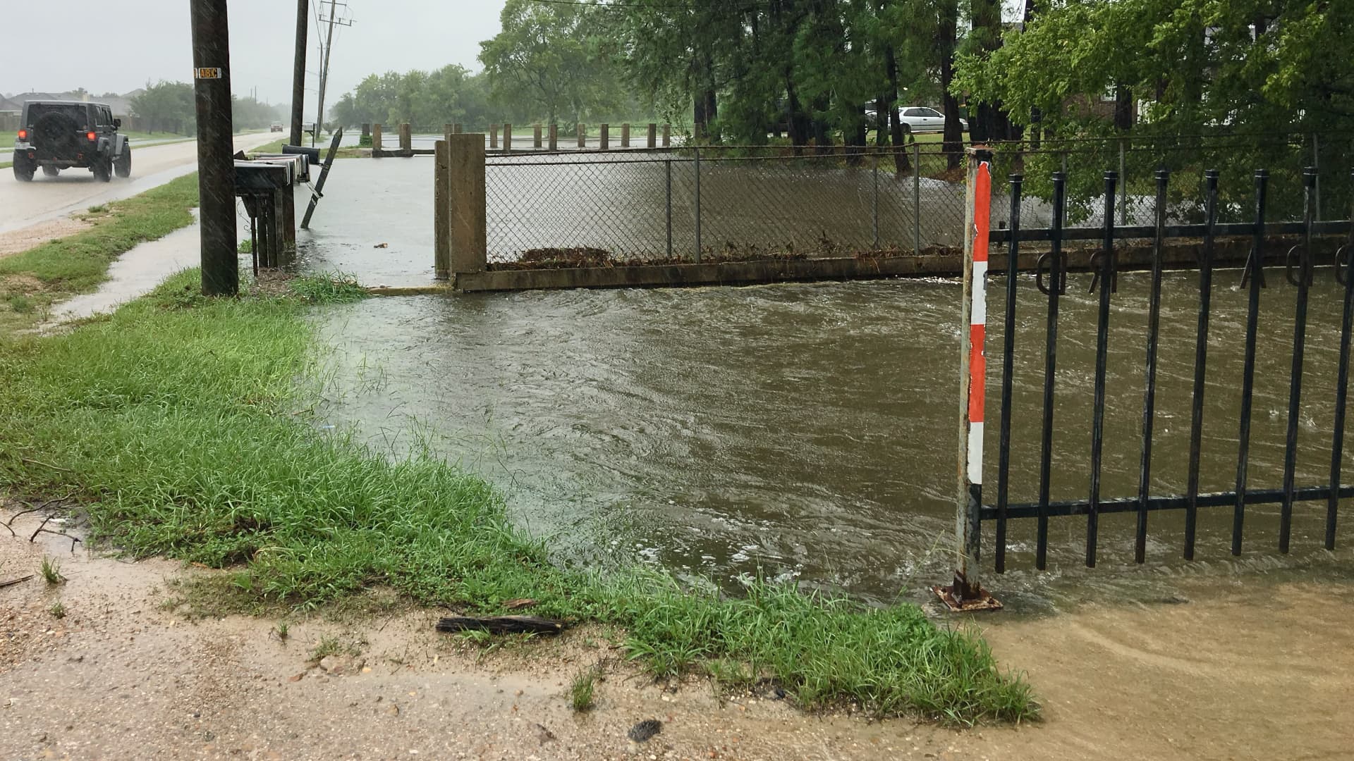 This photograph shows a flooded area with rising water behind a metal gate and fence.