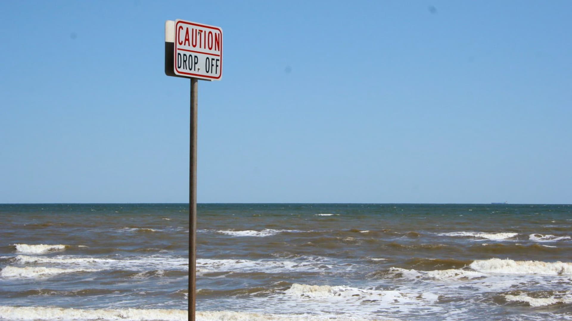 This image shows a "CAUTION DROP OFF" sign standing in the ocean near a sandy beach with waves rolling in.