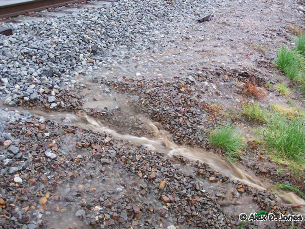 Water is flowing fast across railroad tracks and gravel. The muddy water looks brown and is moving quickly over the rocks and train tracks.