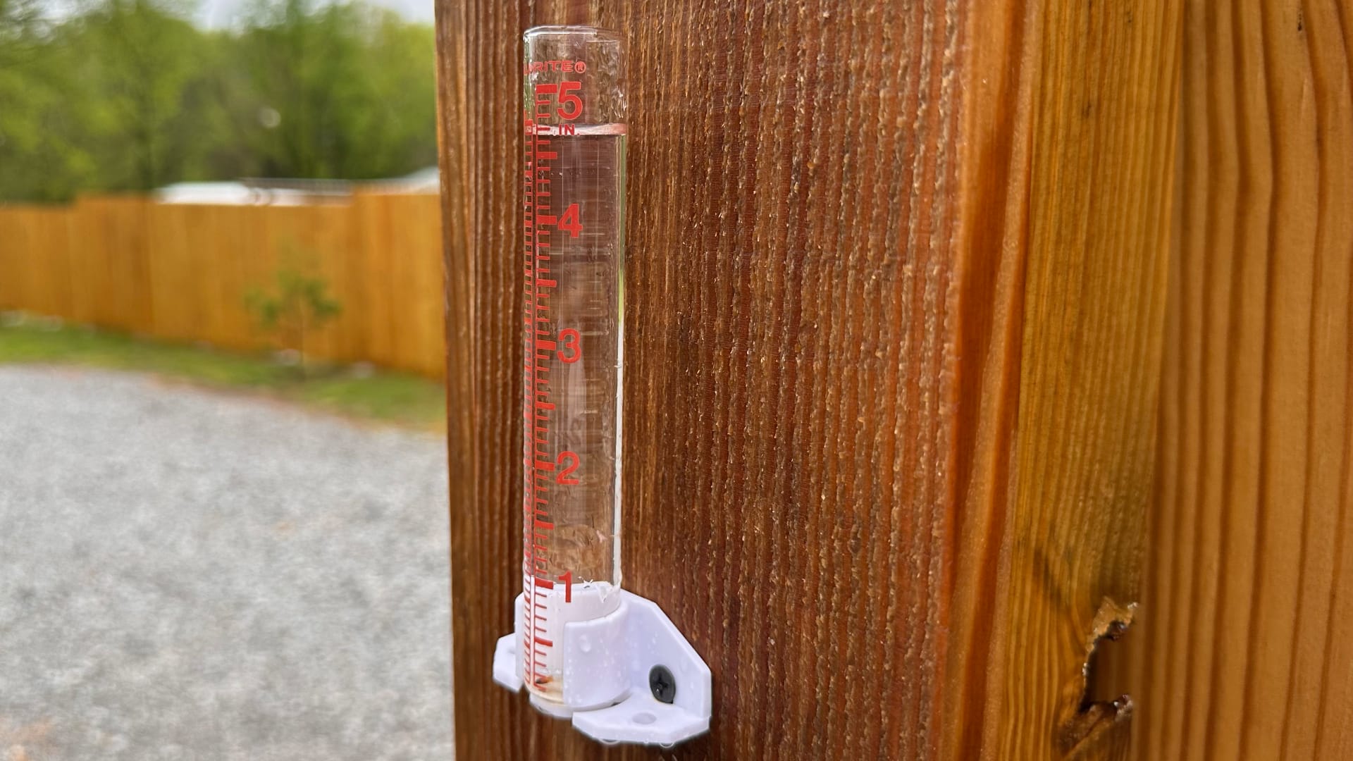 This image shows a rain gauge mounted on a wooden post or fence. A rain gauge is a clear tube with numbers and markings that measure how much rain falls from the sky.