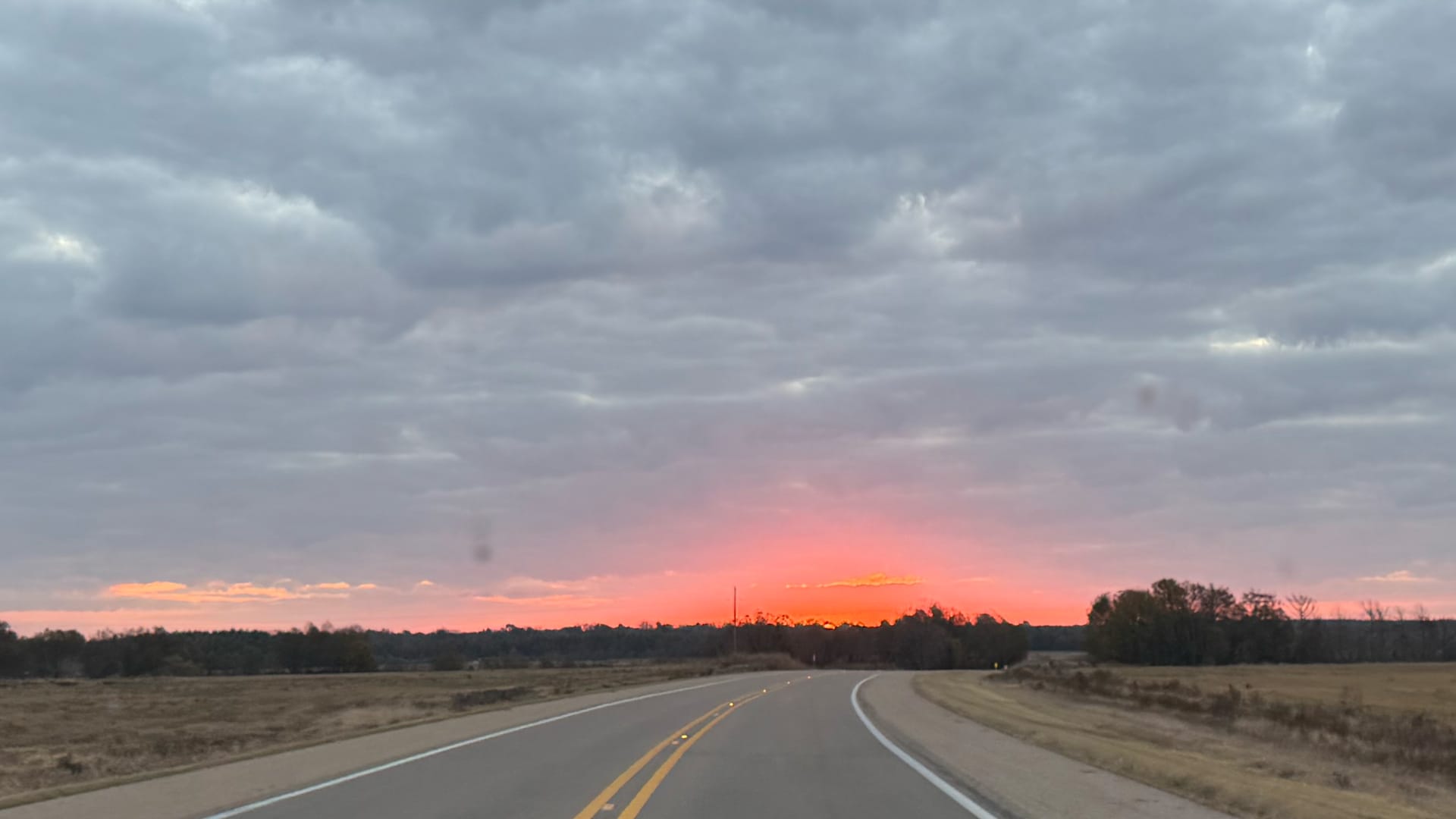 The image shows a road with fields and trees on either side, leading towards a vibrant orange and pink sky.
