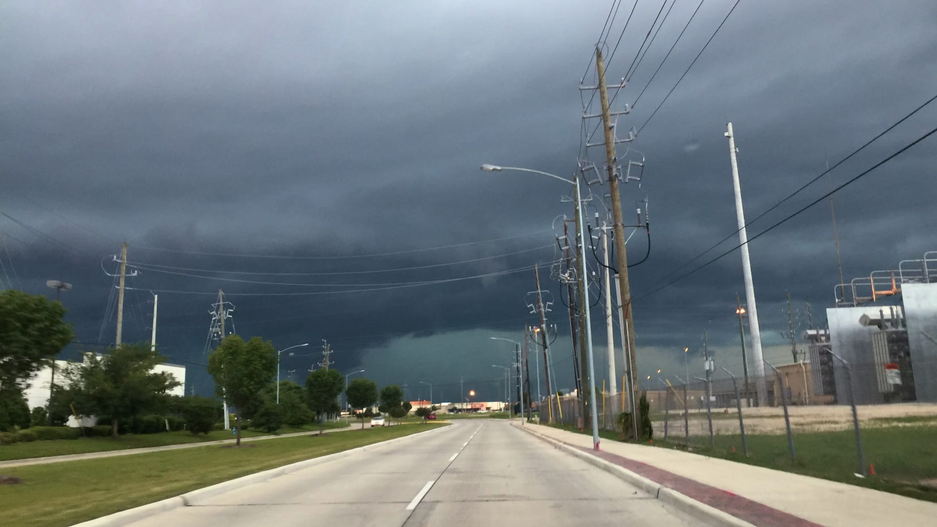 This image shows a dramatic storm moving into a community on a clear day. Dark, heavy clouds are building up over a lake or large body of water in the distance, while the foreground shows streets,...