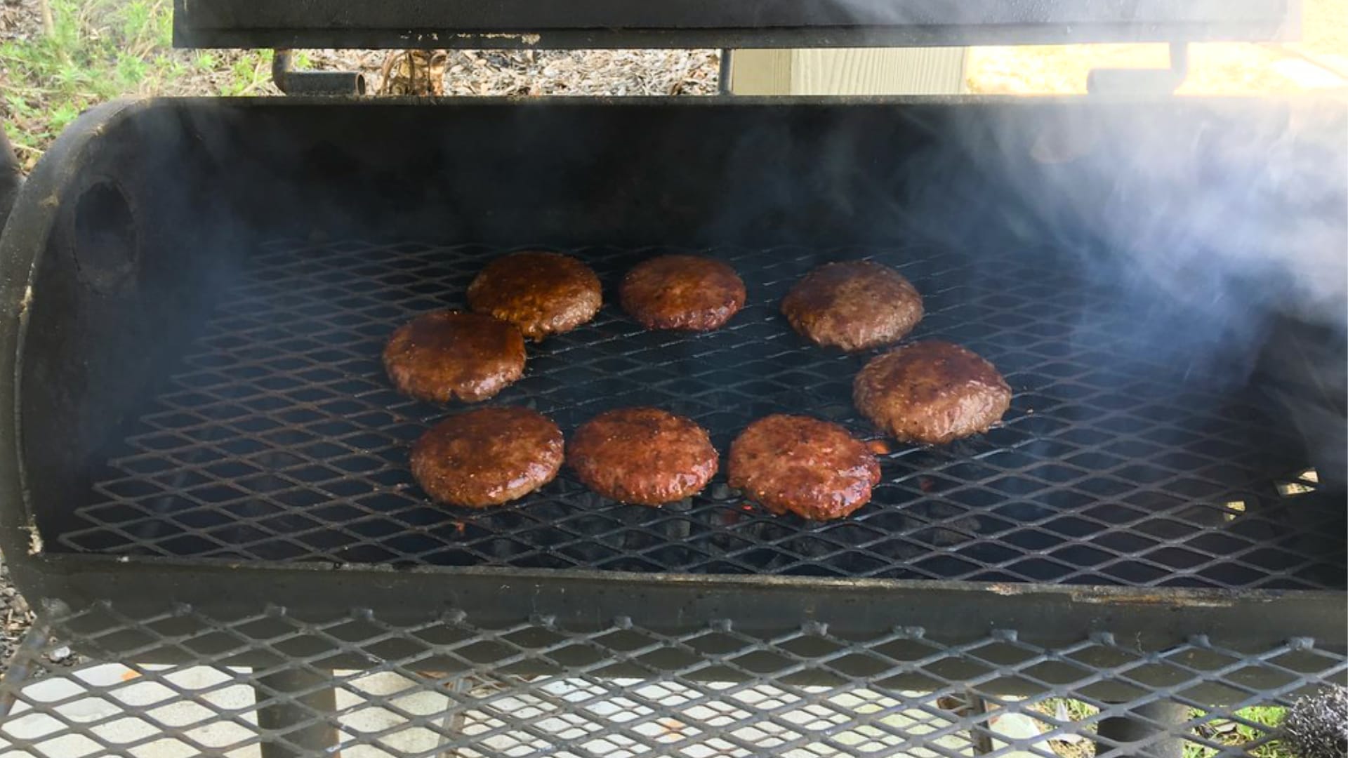 This image shows several hamburger patties cooking on a grill grate inside a black smoker or barbecue.