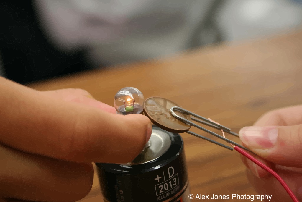A person is using wires to connect a small light bulb to a battery. The light bulb is glowing, which shows that electricity is flowing through the wires from the battery to make the bulb light up.