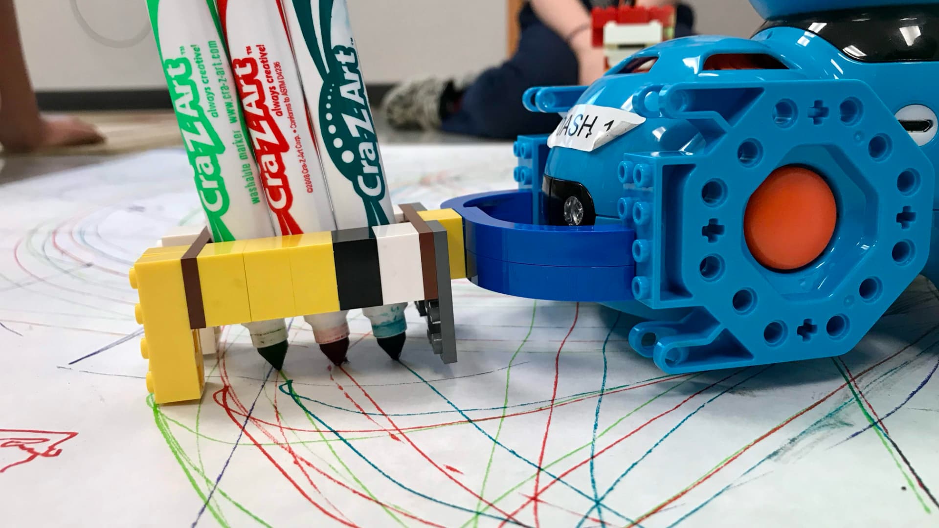 The photo shows a robotic device with markers attached, drawing colorful lines on a large sheet of paper.