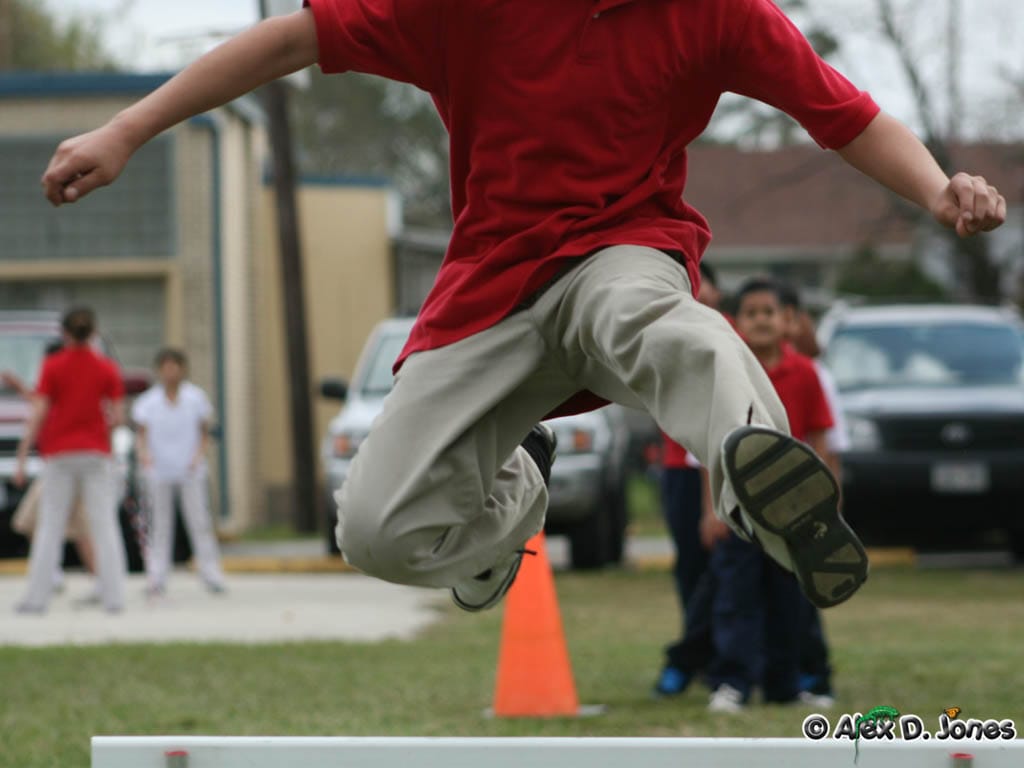 A student wearing a red shirt and gray pants is jumping over an orange cone during an outdoor activity.