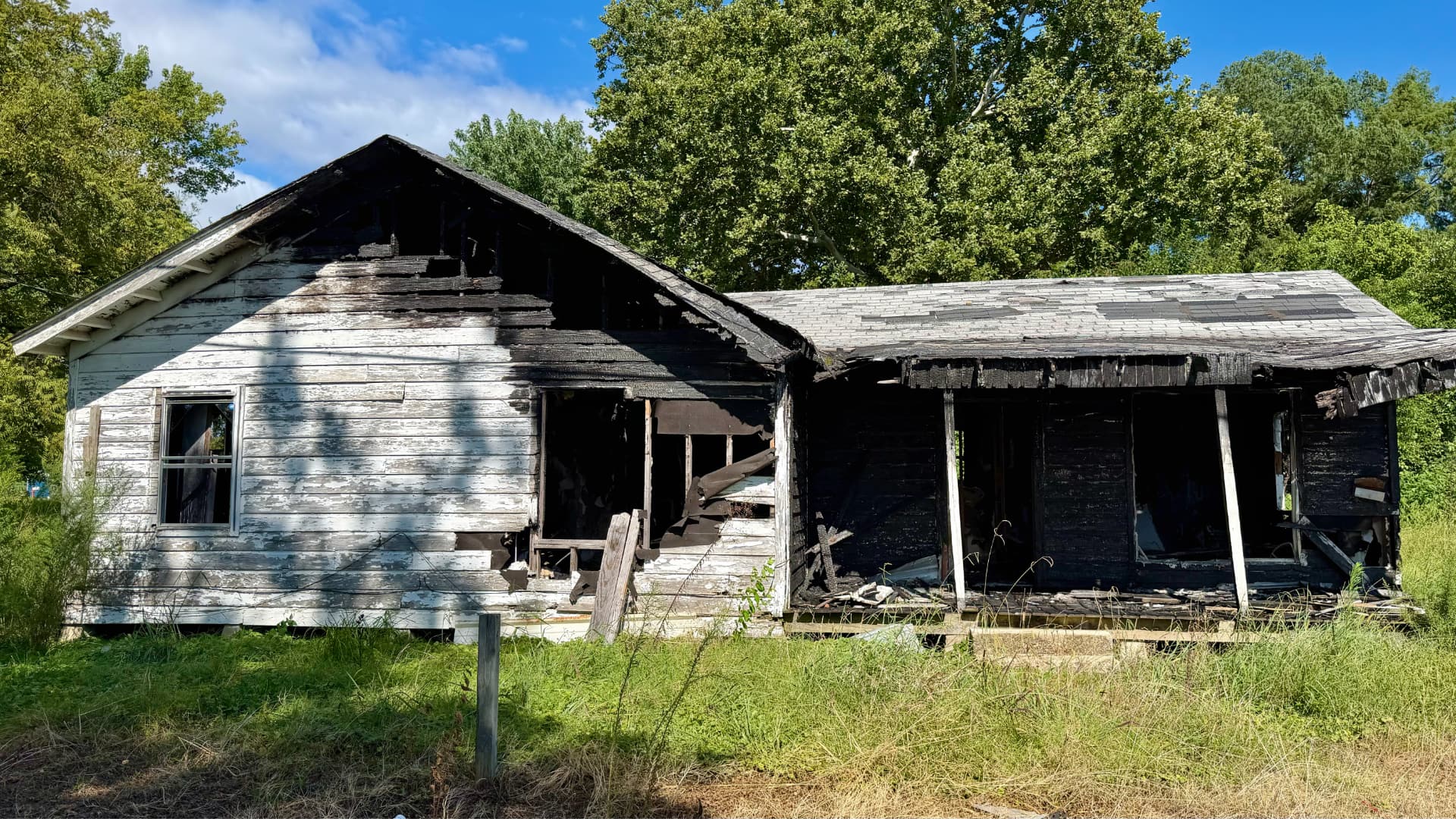 The image shows a house that has been significantly damaged by fire. Large sections of the house are blackened and appear to be burned away, with exposed wooden beams and a collapsed roof section.