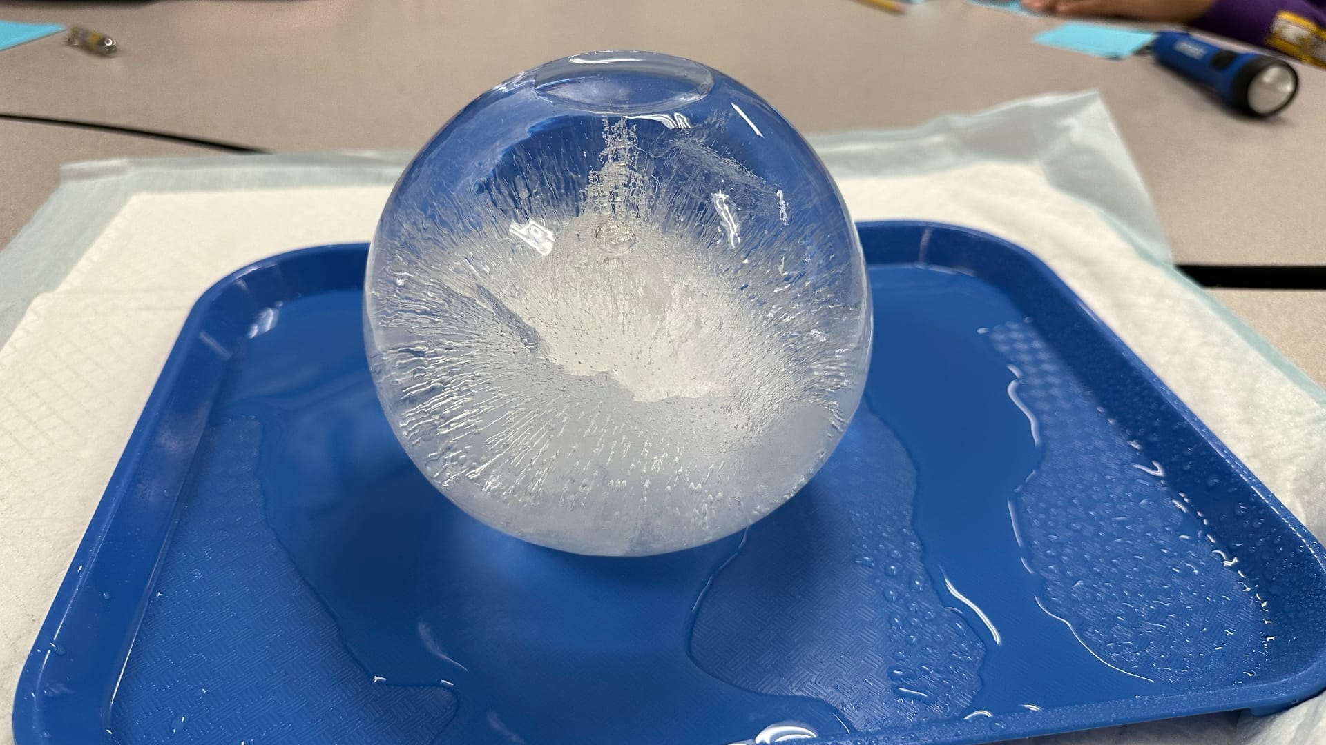 A large, clear ice ball sits in a blue plastic tray. The ice has a cloudy white center that looks like it froze from the outside in.