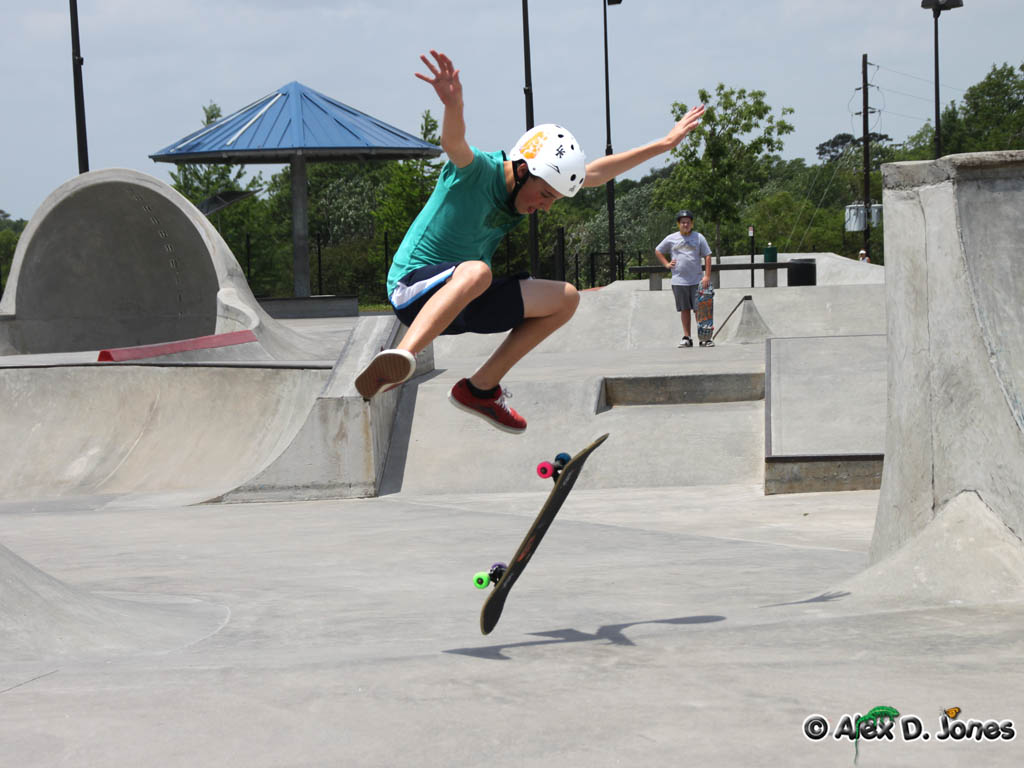 In this image, a skateboarder is performing a trick at a concrete skate park, jumping high into the air while the skateboard flips beneath them.