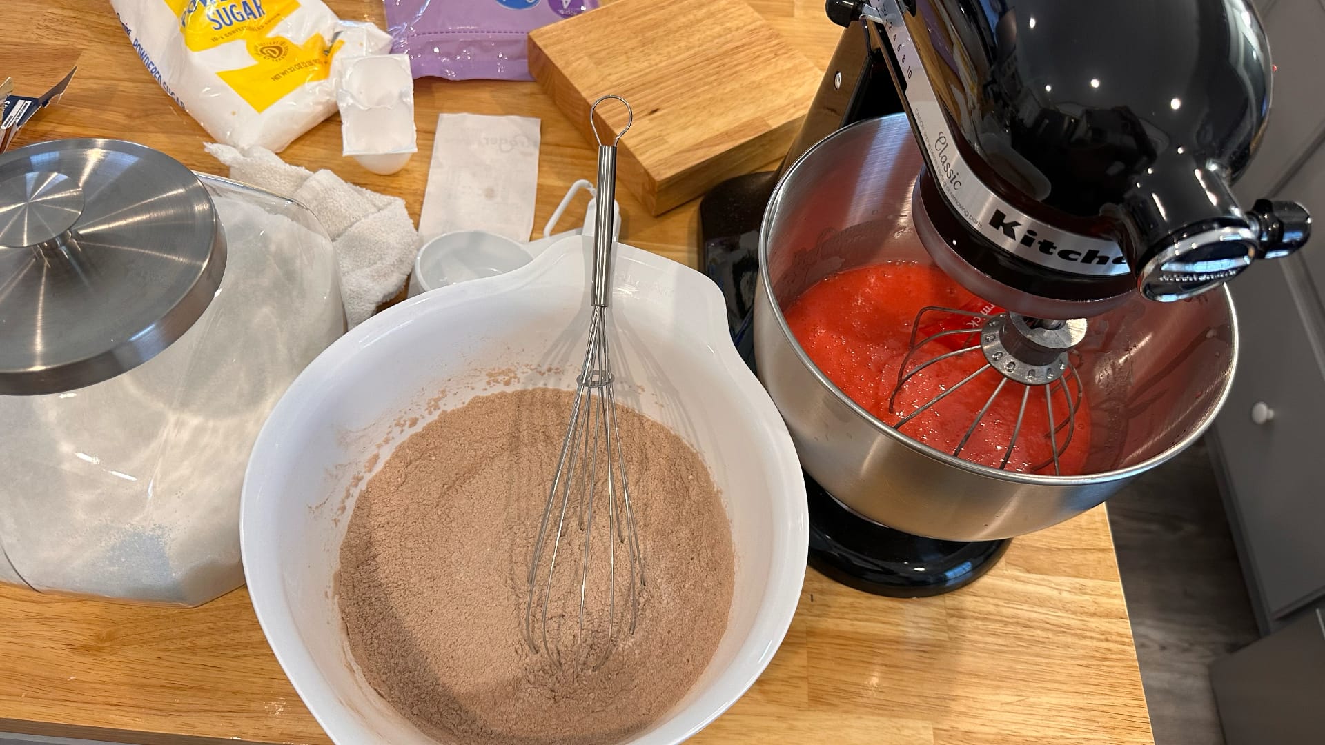 This image shows a cooking scene with dry ingredients in a white bowl (cinnamon and flour mixed together) and a stand mixer containing a red liquid, representing a mixture being created.